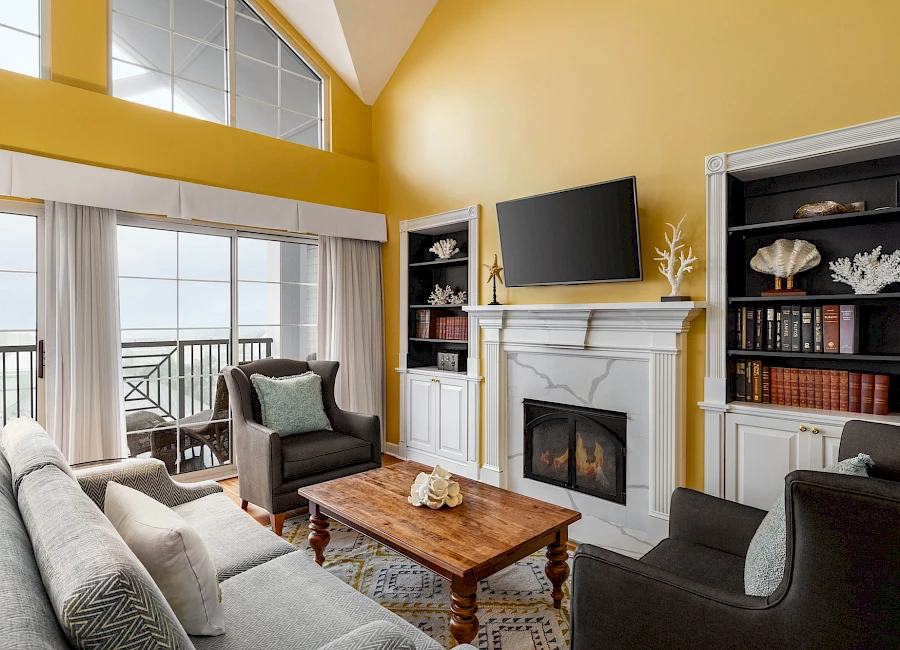 A bright living room with yellow walls, a white fireplace, built-in shelves, a wall-mounted TV, and a cozy seating area facing a coffee table, plus a balcony overlook.
