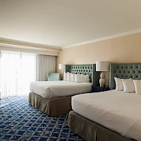 A hotel room with two queen beds, tufted headboards, nightstands, lamps, a chair, and a large window with curtains, plus a dresser—cozy and neat.