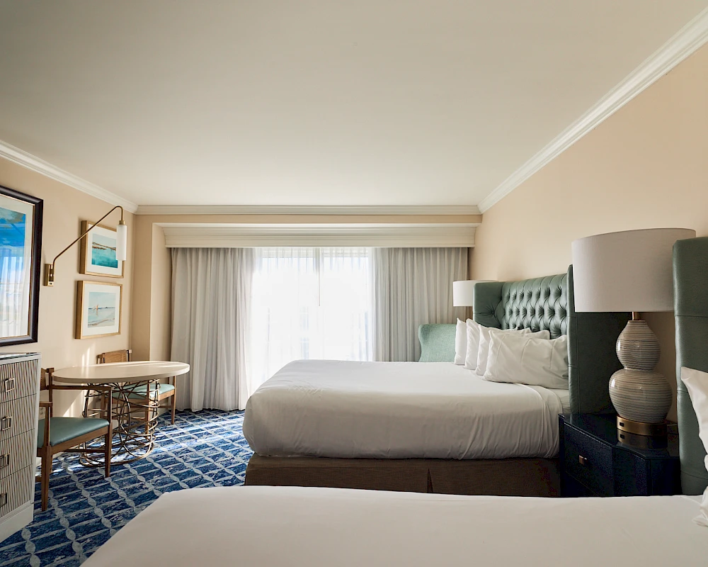 A hotel room with two double beds, a patterned blue carpet, dresser, flat-screen TV, nightstands, and a large window with sheer curtains ending with a view.