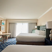 A hotel room with two double beds, a patterned blue carpet, dresser, flat-screen TV, nightstands, and a large window with sheer curtains ending with a view.