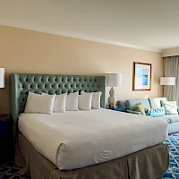 A hotel room with a large bed, green tufted headboard, two bedside lamps, a seating area with blue-green sofa, and a windowed balcony.