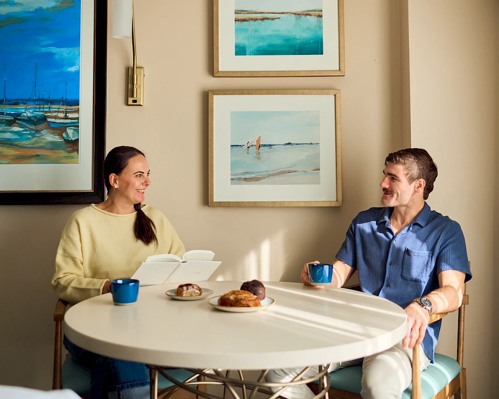 Two people sit at a round table chatting over coffee and pastries in a cozy cafe adorned with framed ocean art.