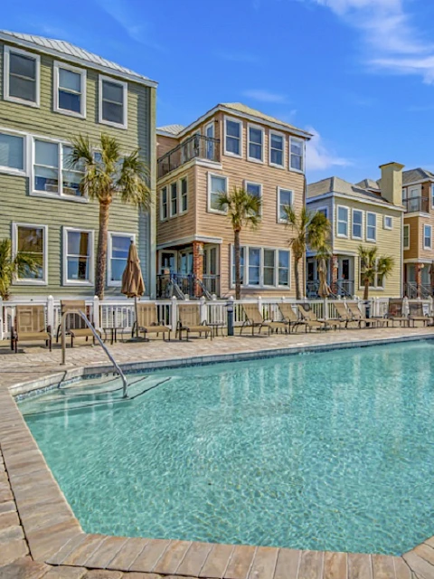 A swimming pool surrounded by colorful three-story houses and palm trees under a clear blue sky.