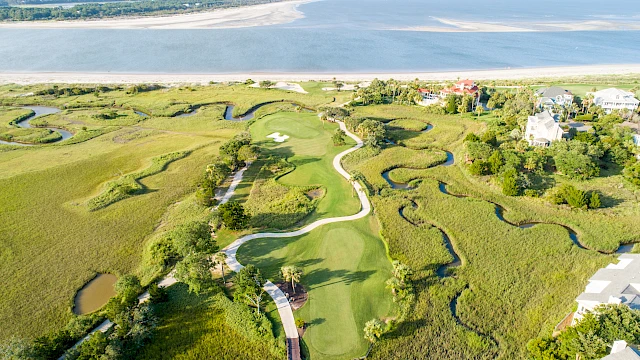 The image shows an aerial view of a coastal golf course with winding paths and surrounding grassy marshlands, near a body of water.
