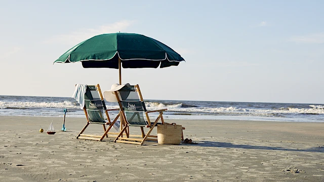 Two beach chairs under a green umbrella facing the ocean, with a straw hat and beach bag nearby on the sand.