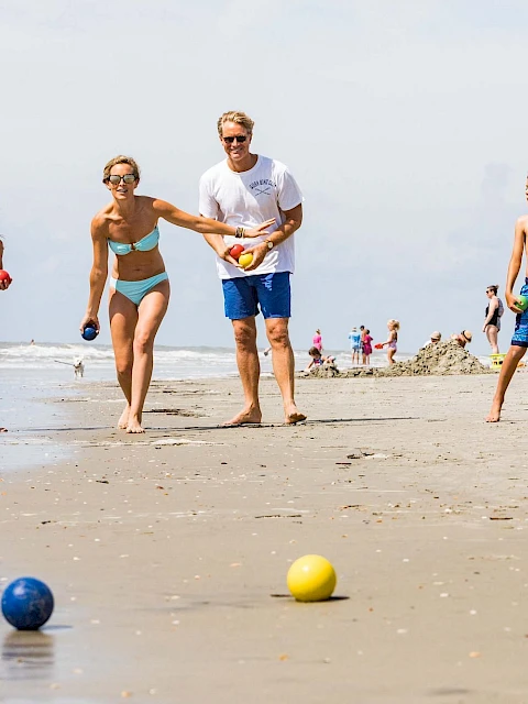 A family is playing bocce on a sandy beach near the ocean, surrounded by people enjoying the sun and beach umbrellas.
