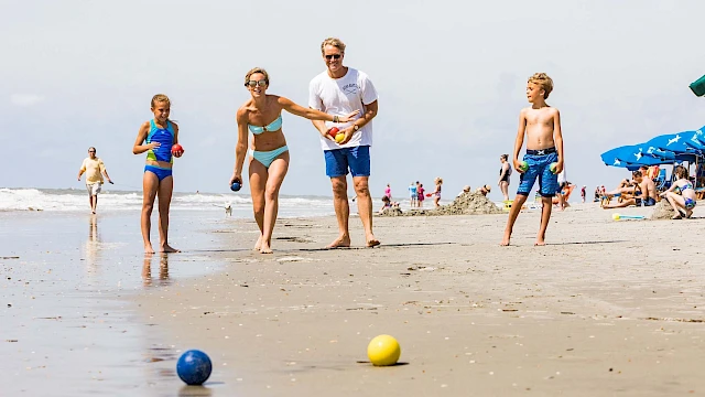 A family is playing bocce on a sandy beach near the ocean, surrounded by people enjoying the sun and beach umbrellas.