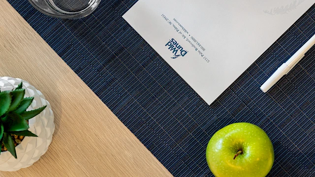 A green apple, pen, glass, paper with "Wild Dunes" logo, and a plant on a wooden desk with a blue mat.