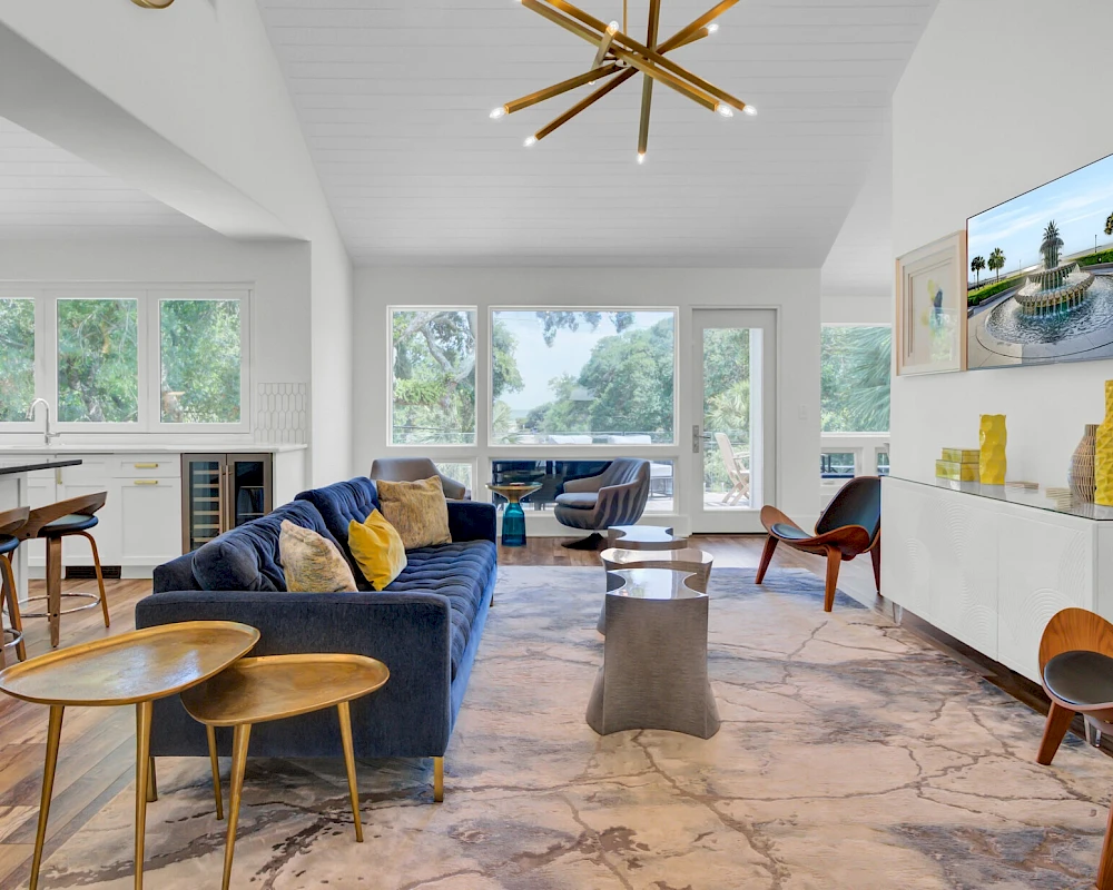 A modern, airy living room with a blue sectional sofa, round wooden coffee tables, floor-to-ceiling windows, a wall-mounted TV, and marble floors, bright and inviting.