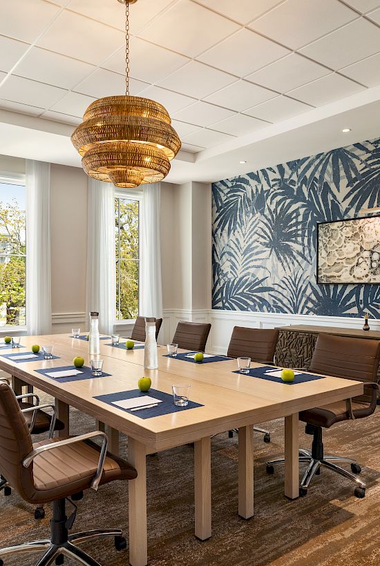 A modern conference room with a wooden table, leather chairs, large windows, and a tropical wallpaper accent wall under a chandelier.