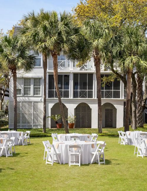 An outdoor event setup with round tables and white chairs on a grassy lawn, surrounded by palm trees and buildings.