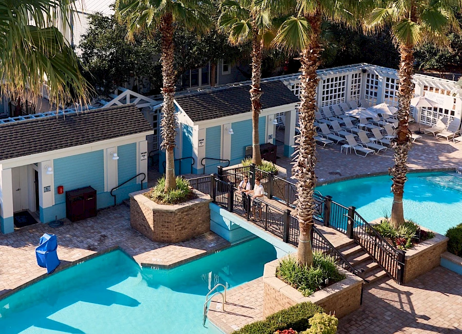 A sunny pool area with turquoise water, palm trees, blue and white buildings, a lounging chair, and stepping platforms around the pool, all under a clear sky.