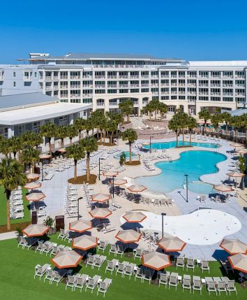 A resort pool area with lounge chairs, umbrellas, and nearby buildings surrounded by palm trees under a clear blue sky.