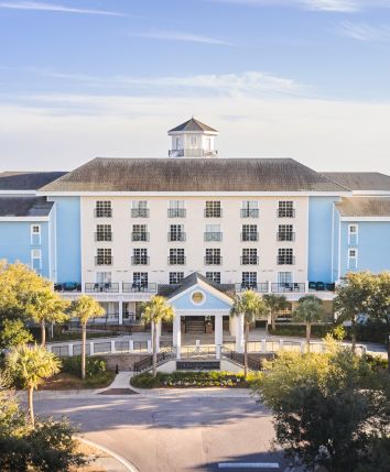 A blue and white multi-story building with a central entrance, surrounded by trees and set near the coast under a clear sky.