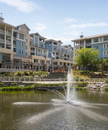 The image shows a modern residential complex with a fountain in a pond in the foreground.