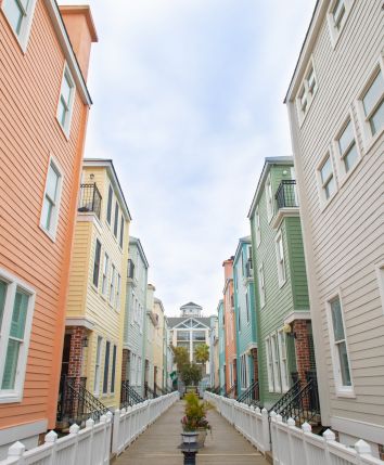 A narrow alleyway with colorful, pastel-colored, three-story houses on either side and a white picket fence along the path.
