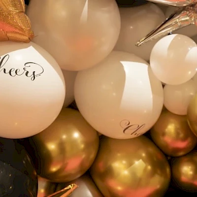 A festive cluster of white, gold, and rose balloons with cursive “Cheers” text, creating a celebratory party vibe.