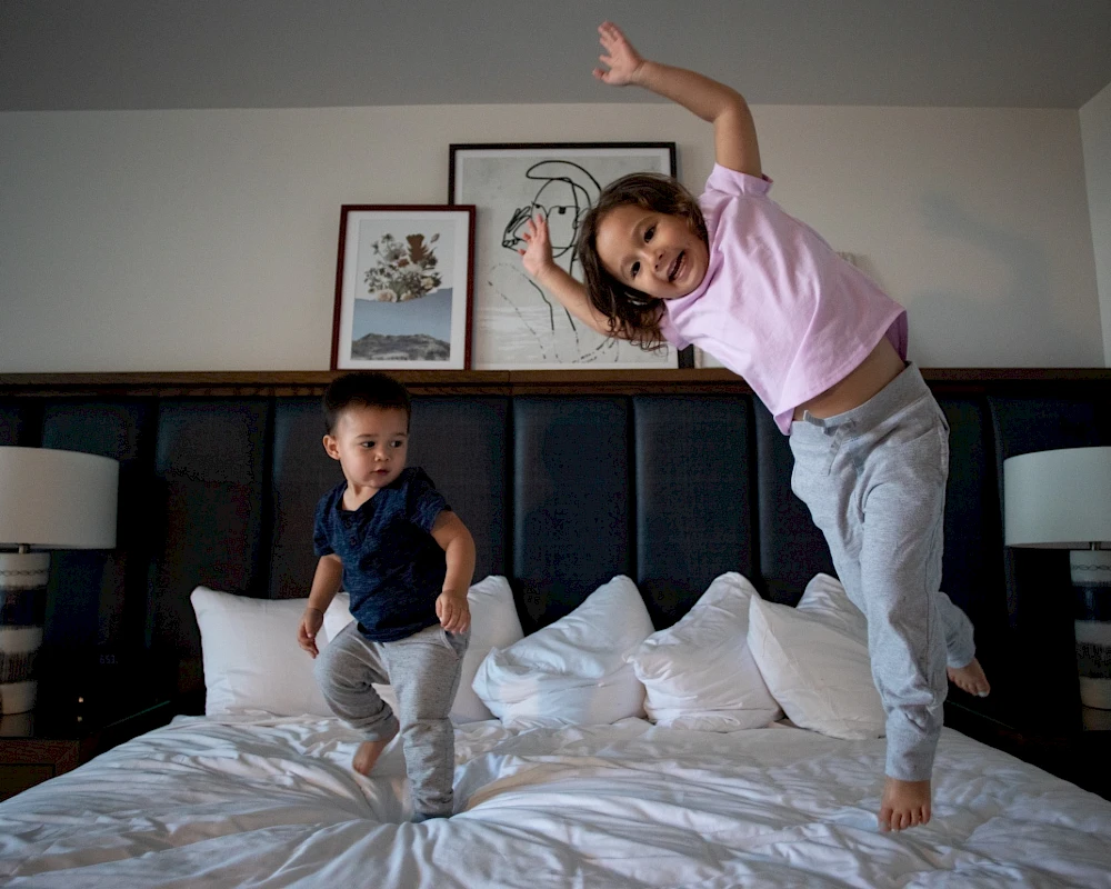 Two kids jump and play on a bed, one mid-air and one landing, in a cozy bedroom with framed wall art behind them.