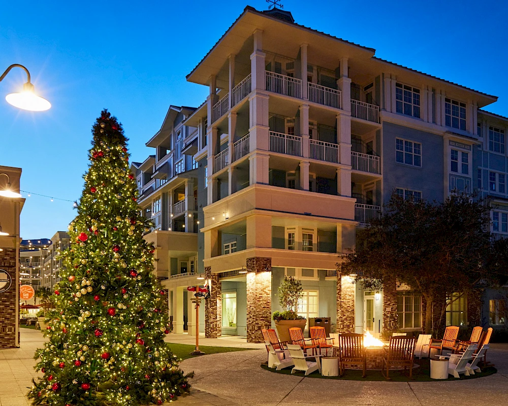 A festive outdoor scene with a tall Christmas tree lit with ornaments beside a modern apartment building and cozy seating around a firepit, at dusk.