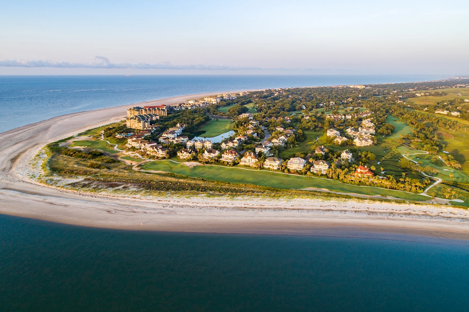 Aerial view of a coastal resort with sandy beaches, turquoise water, and a cluster of white buildings/houses surrounded by green landscaping along a curved shoreline.