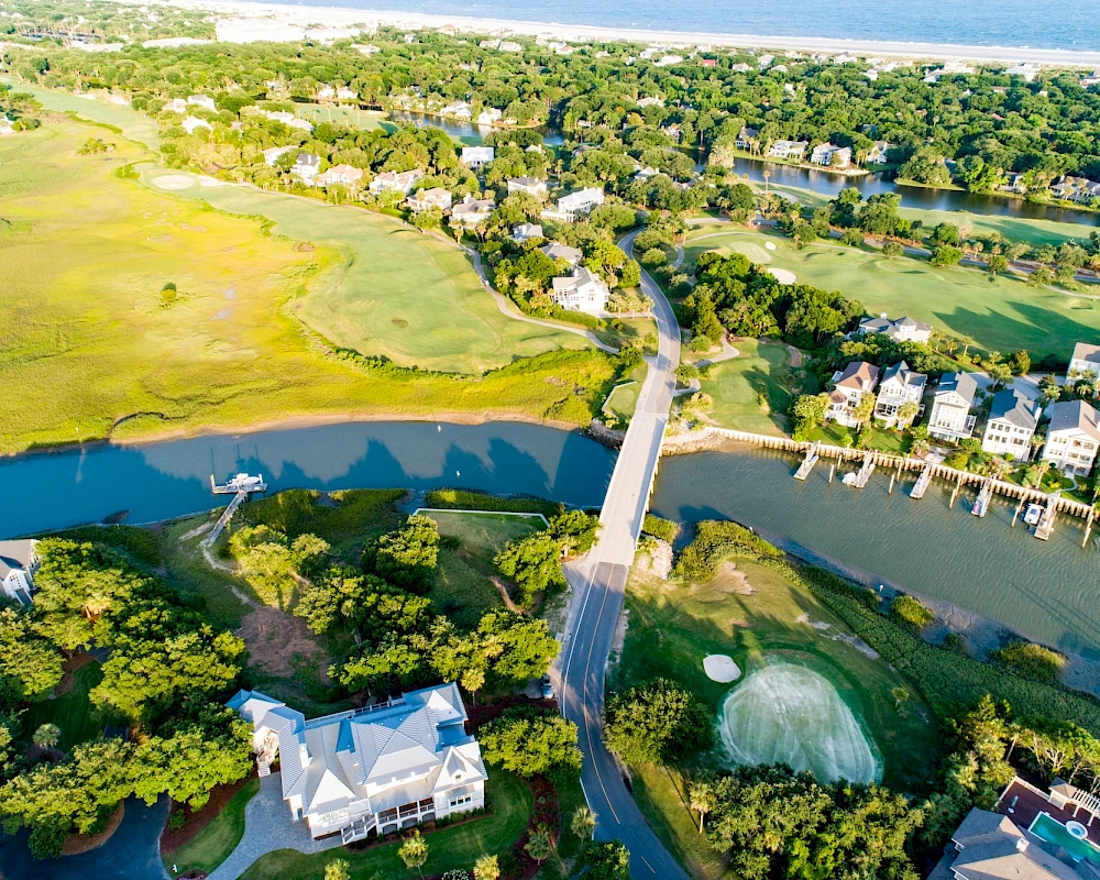 Aerial view of a coastal suburb with a bridge over water, winding roads, houses, lush greenery, and a sandy marshy area near the coast.