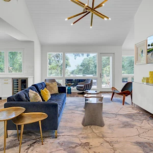 A bright, airy living room with a blue sofa, mid-century chairs, marble floor, large windows, and a wall-mounted TV display; stylish decor and natural light.