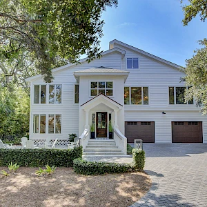 A large, white two-story house with a peaked entry porch, multiple windows, and a three-car garage, surrounded by tall trees and a paved driveway.