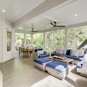 A bright, airy screened porch with white walls, blue cushioned sectional sofa, wicker chairs, ceiling fans, and a dining area overlooking lush greenery.