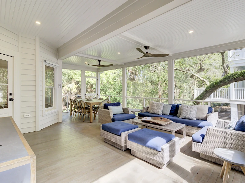 A bright, airy screened porch with white walls, blue cushioned sectional sofa, wicker chairs, ceiling fans, and a dining area overlooking lush greenery.