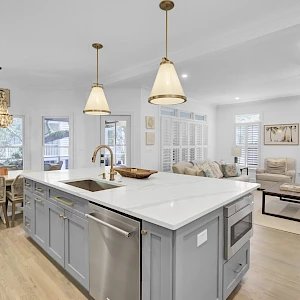 Modern open-concept kitchen and living area with a large island, light gray cabinetry, pendant lights, and light wood flooring.