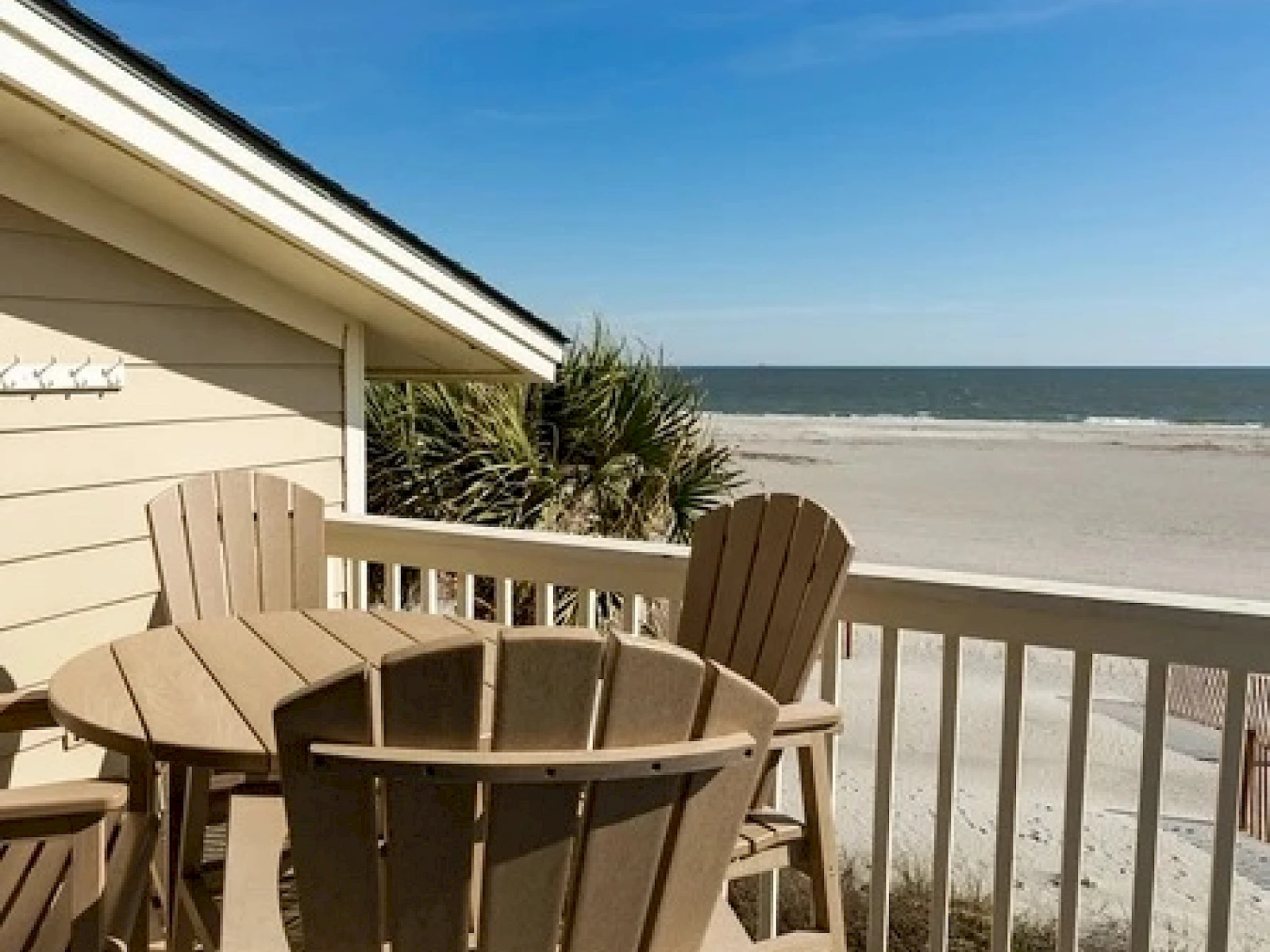 A sunny beach view with a wooden deck and beige Adirondack chairs around a round table overlooking the sandy shore and blue ocean waves.