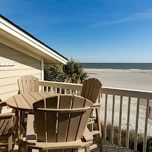 A sunny beach view with a wooden deck and beige Adirondack chairs around a round table overlooking the sandy shore and blue ocean waves.