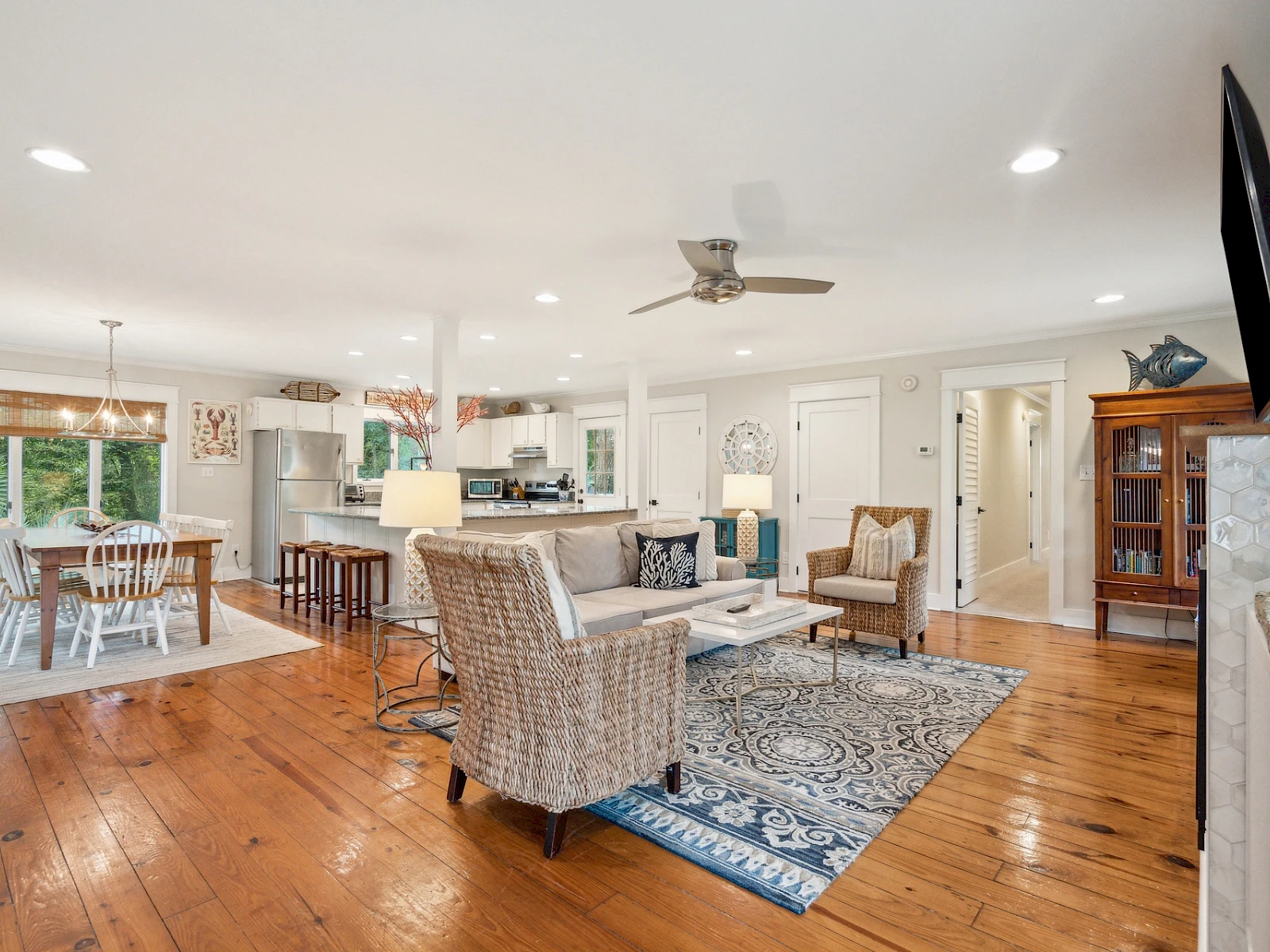 Spacious open-plan living room and kitchen with hardwood floors, neutral furniture, a blue rug, large windows, and a TV mounted above a fireplace.