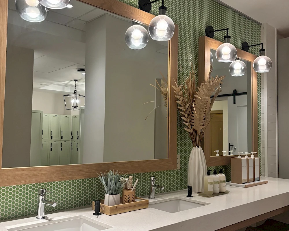 A modern bathroom vanity area with two large mirrors, green tiled backsplash, multiple light fixtures, and neatly arranged accessories on the counter.