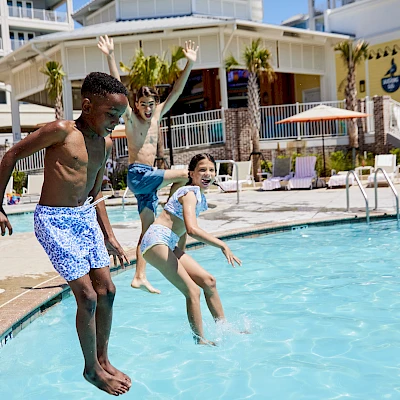 Three kids jump into a sunny hotel pool, splashing joyfully as others relax nearby under umbrellas. Always ending the sentence.