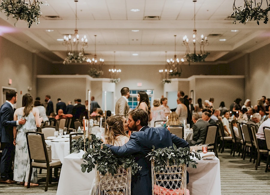A wedding reception: a couple sits at the head table, guests mingle and toast in a warmly lit banquet hall. 140 characters, ending with a period.