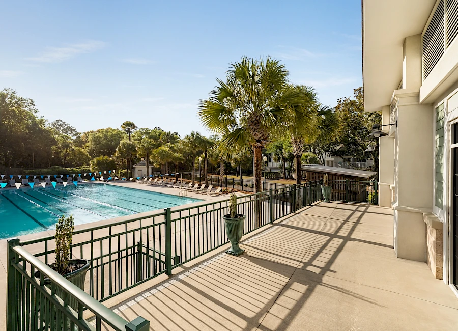 A sunny outdoor pool area with clear blue water, palm trees, lounge space, and a balcony railing overlooking the pool.