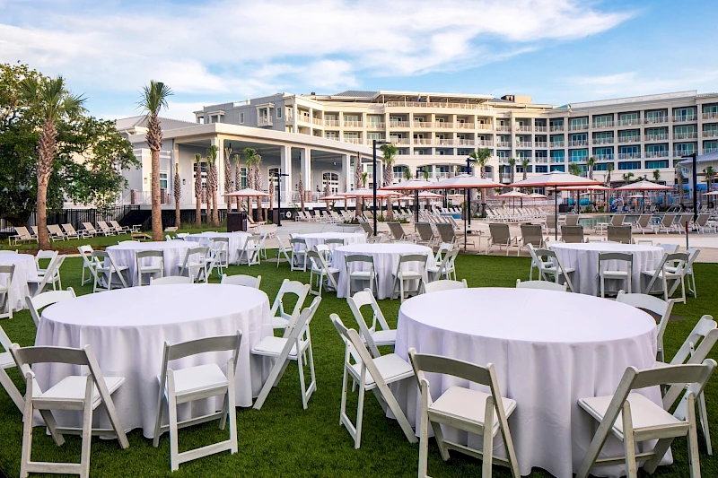 An outdoor event setup with round tables draped in white cloths and white folding chairs on a grassy lawn, near a resort-style building.