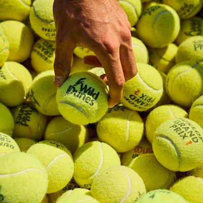 A hand picks a bright yellow tennis ball from a pile of many similar balls.