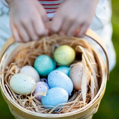 A basket of colorful speckled Easter eggs nestled in straw, held by hands over a grassy background.